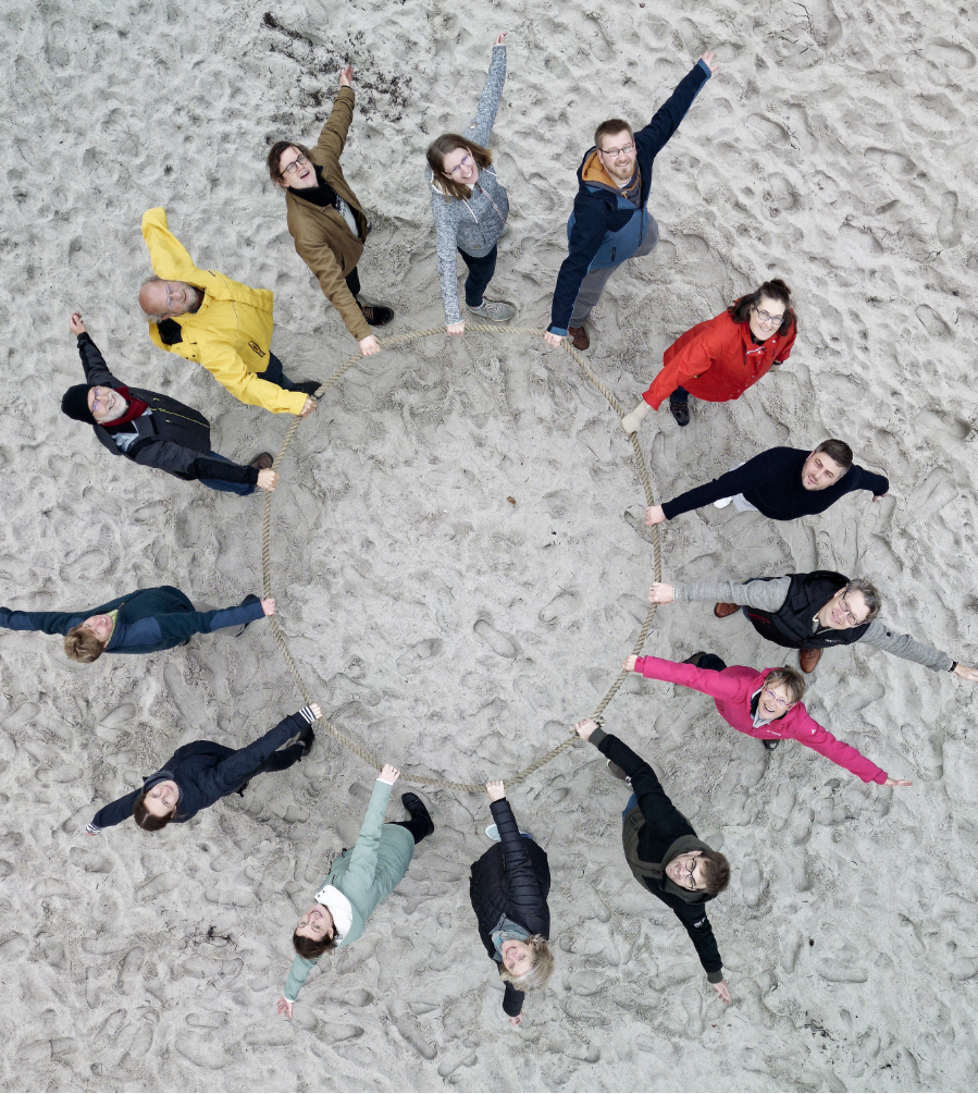 eam von Mitarbeitenden im Kreis am Strand mit Seil aus der Vogelperspektive